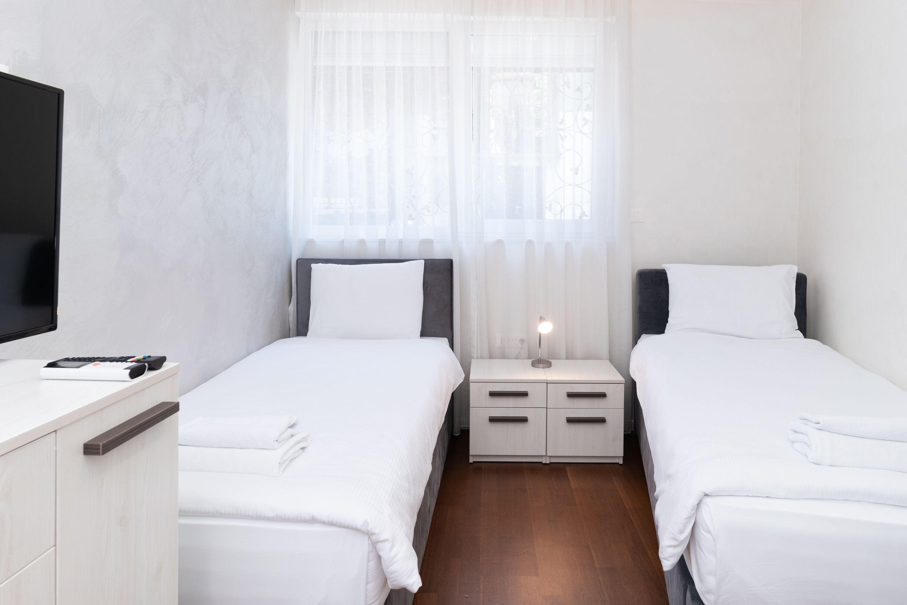 Une chambre d’hôtel avec deux lits simples, une table de chevet avec une lampe, un téléviseur sur le côté gauche et un plancher en bois.