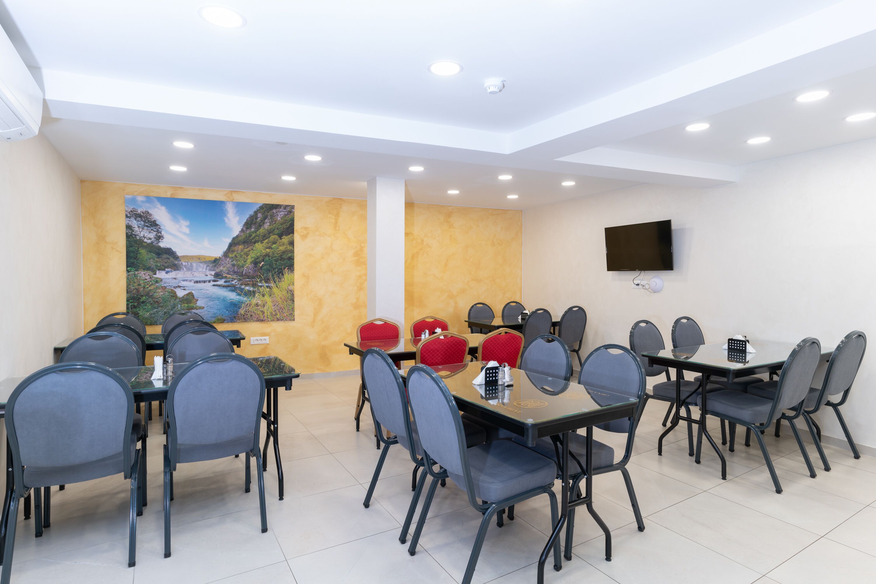 A conference room with multiple tables and chairs, a large painting of a landscape, and a mounted TV on the wall.