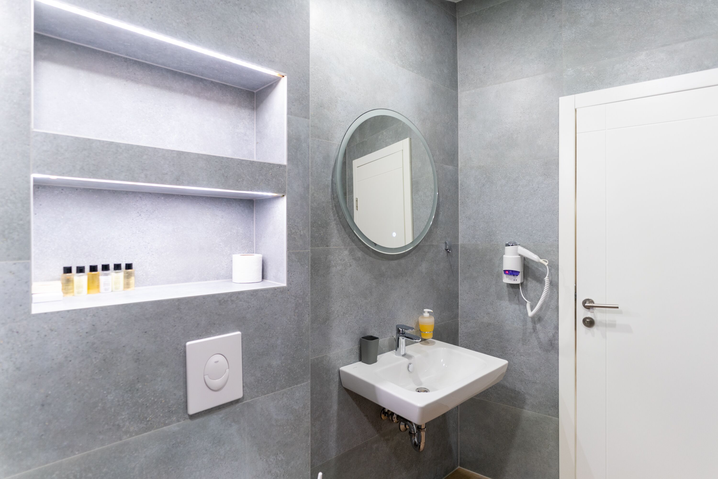 A modern bathroom with a white sink, a round mirror, and a hairdryer mounted on the wall.