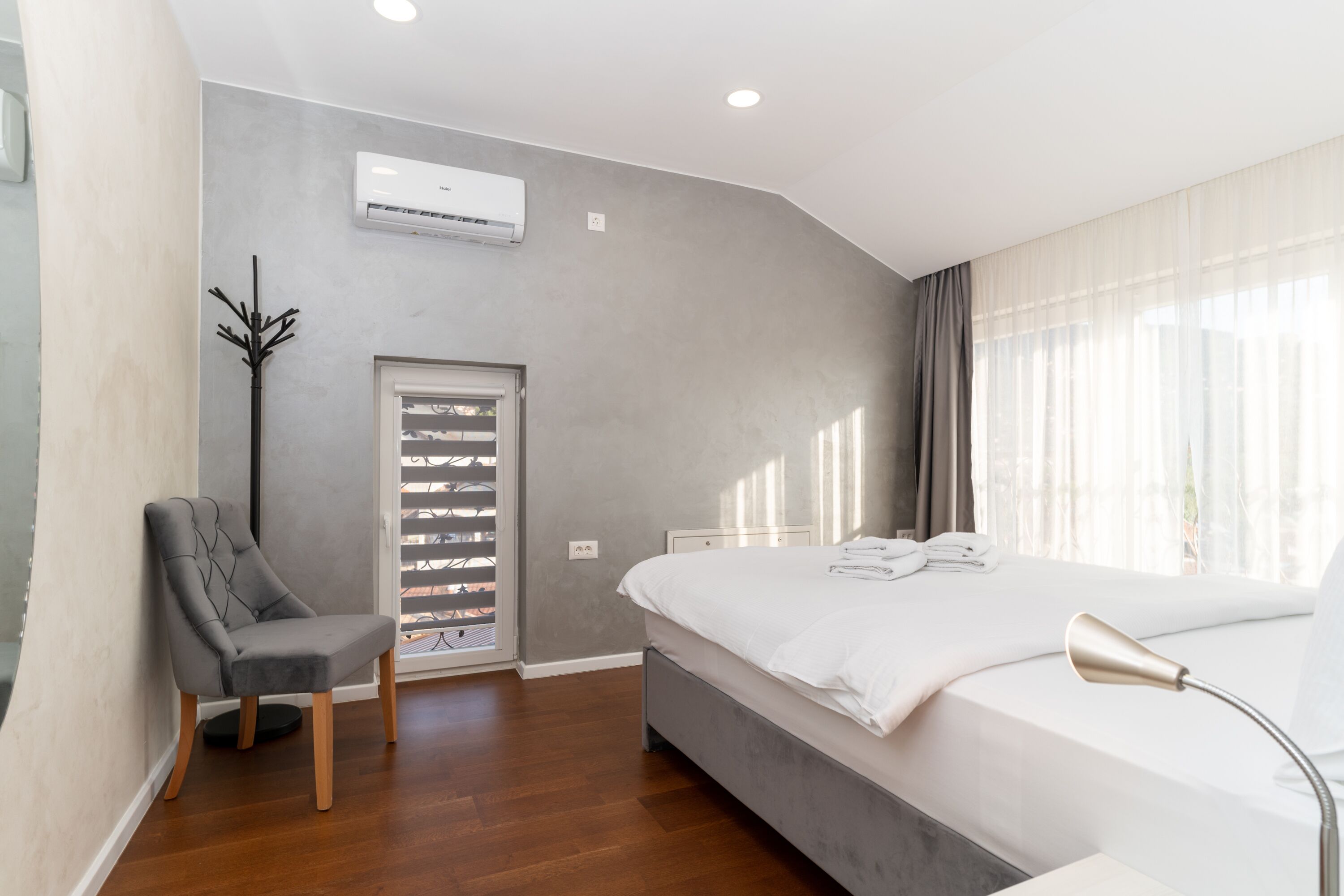 A modern hotel room with a large bed, a chair, a window with curtains, and an air conditioning unit.