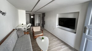 A modern living room with a sofa, a small table, and a chair. There is a TV mounted on the wall and a door on the right.