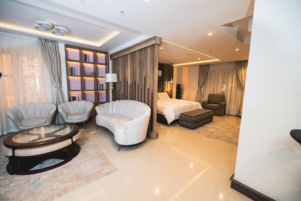 A modern hotel room with a curved sofa, a round coffee table, a bed, and a seating area with two armchairs.