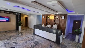 A hotel reception area with a marble floor, a reception desk, a TV screen displaying news, and a wooden reception desk with a clock and a flower arrangement. - HABAB GRAND RESORT (Abuja)