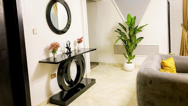 A modern living room with a grey sofa, a black console table, a large potted plant, and a mirror on the wall. - HABAB GRAND RESORT (Abuja)