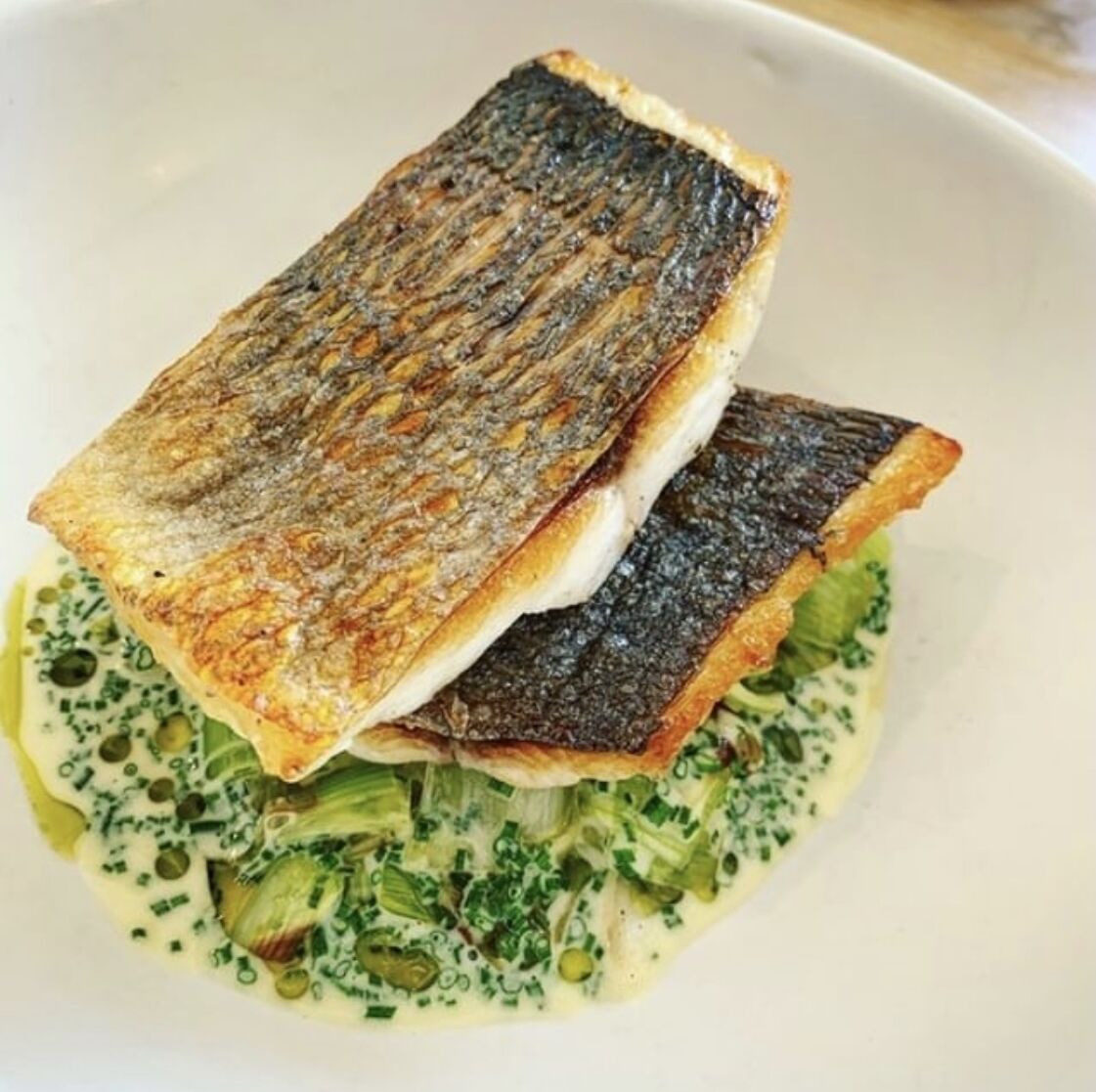 A white plate with a piece of grilled fish, a creamy sauce, and chopped green herbs.