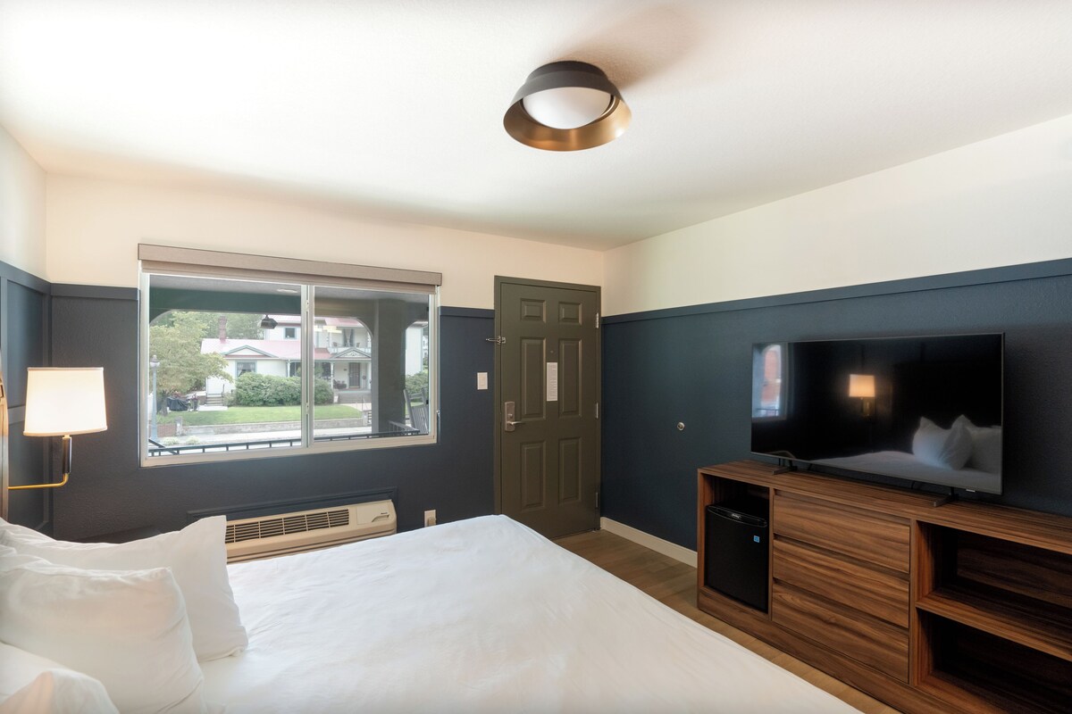 A hotel room with a bed, a television, a window with a view of buildings, and a door.