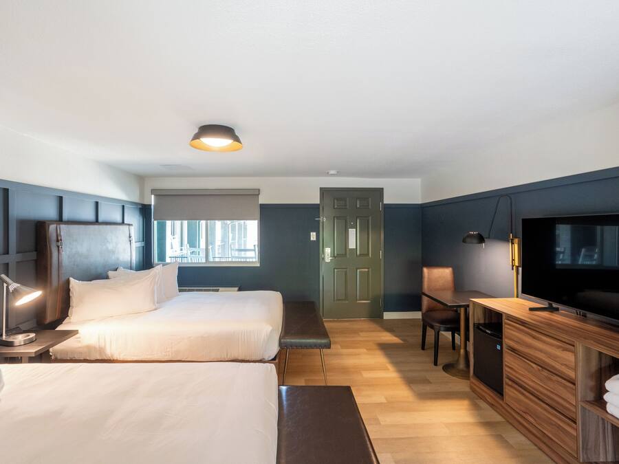 A hotel room with two beds, a desk, a television, and a bench.