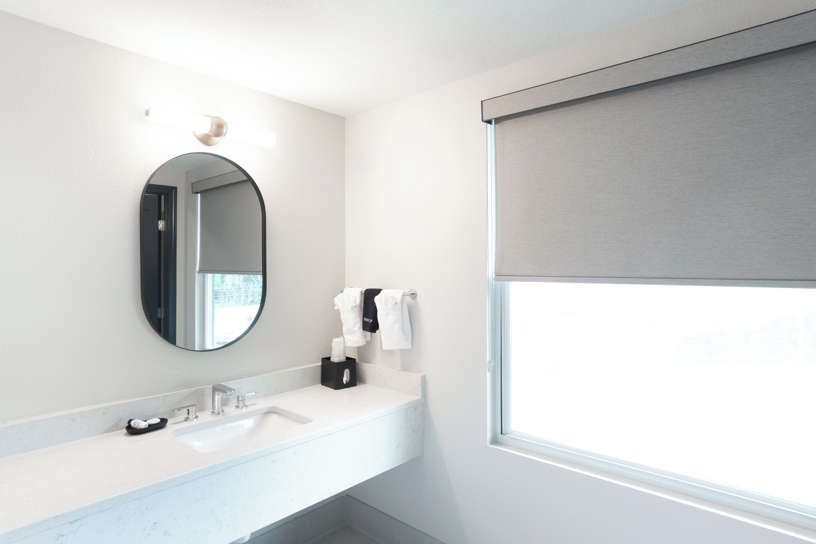 Une salle de bain moderne avec un lavabo blanc, un miroir rond et une fenêtre avec un store.