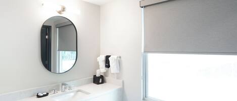 A modern bathroom with a white sink, a round mirror, and a window with a blind.