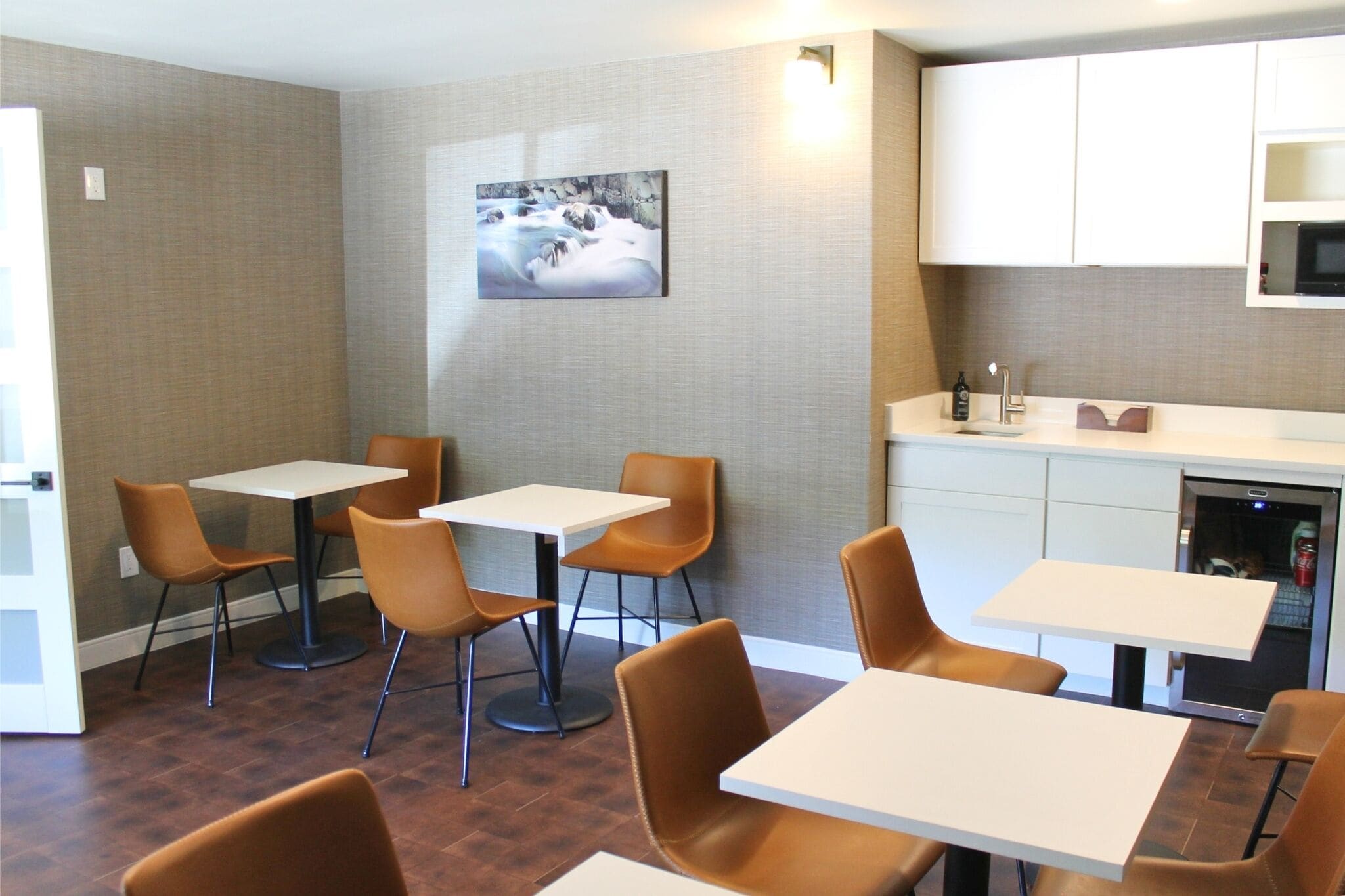 A dining area with tables and chairs, a kitchen area with cabinets and a microwave, and a painting on the wall.