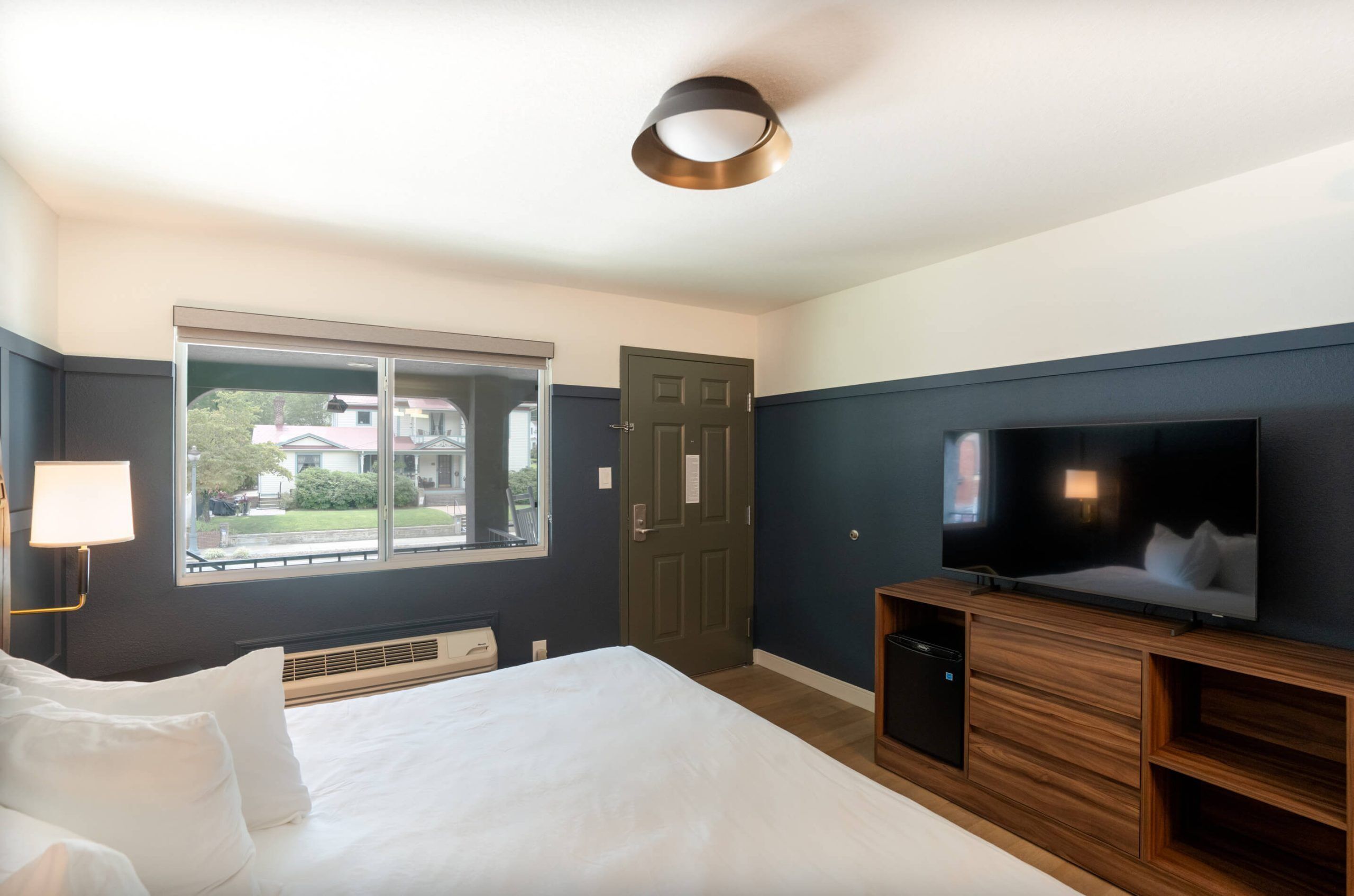A hotel room with a bed, a TV, a window with a view of buildings, a door, and a lamp.