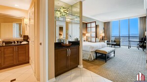Habitación de hotel con una cama grande, zona de estar con vistas y baño con tocador y espejo.