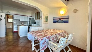 Un coin repas avec une table recouverte d’une nappe à motifs, des chaises blanches et une cuisine en arrière-plan.