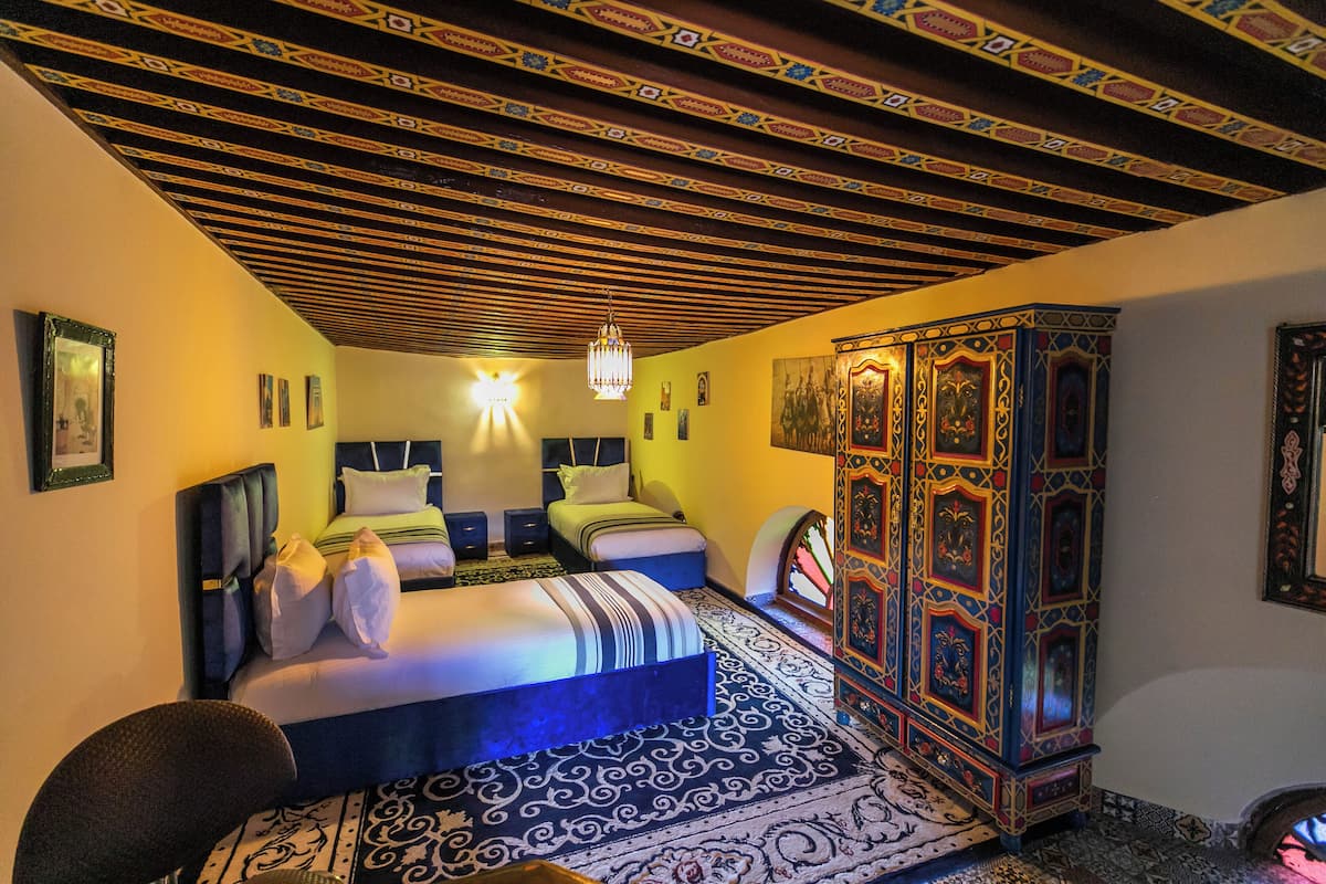 A room with a bed, two bedside tables, a chandelier, a decorative cabinet, and patterned rugs.