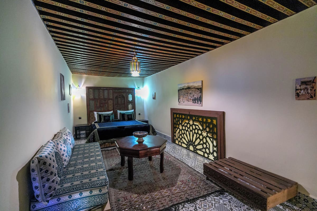 A room with a patterned sofa, a wooden table, and a bench. There is a bed with a headboard in the background.