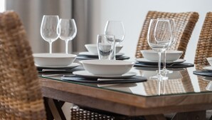 A dining table set with white plates, bowls, and clear wine glasses, surrounded by wicker chairs.
