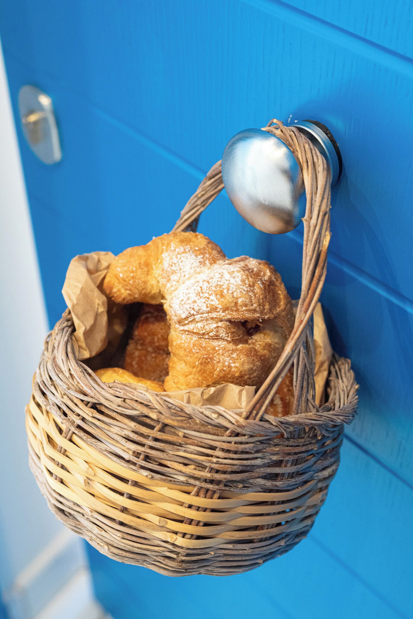 Un panier en osier rempli de pâtisseries, accroché à une porte bleue.
