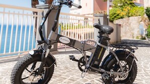 En stationær elcykel parkeret på brosten med gelænder og træer i baggrunden.