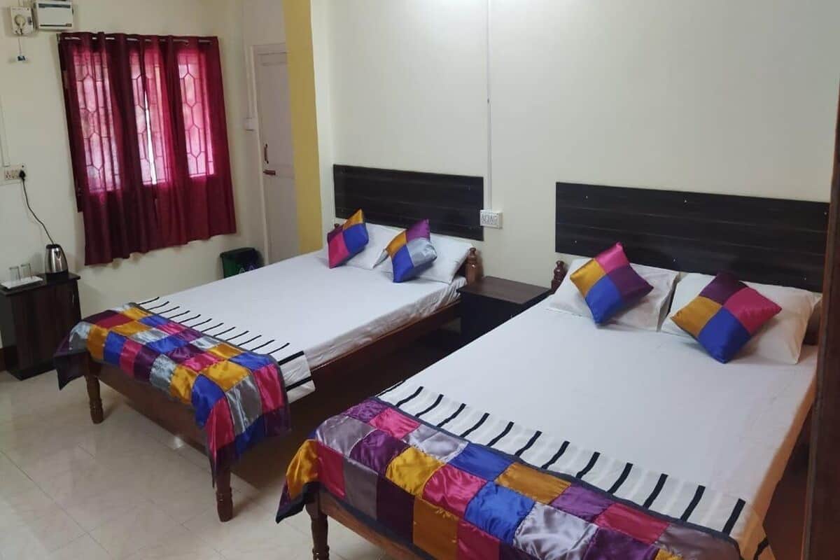 Quarto de hotel com duas camas de solteiro, cada uma com um edredom colorido, uma cabeceira de madeira e uma parede branca.