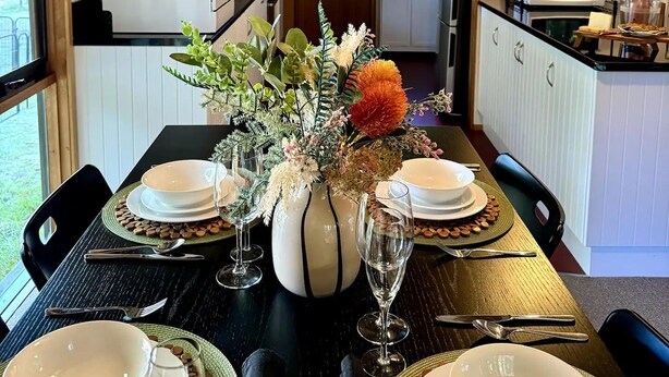 Ein Essbereich mit Holzbalkendecke, einer Küche im Hintergrund und einem Tisch für vier Personen, gedeckt mit Tellern, Gläsern und einer Blumenvase.