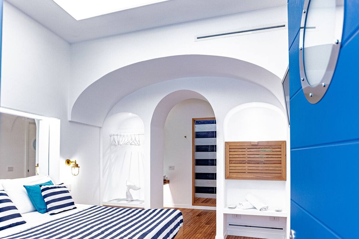 A modern bedroom with a wooden floor, a bed with striped bedding, a white arched doorway, and a blue accent wall.