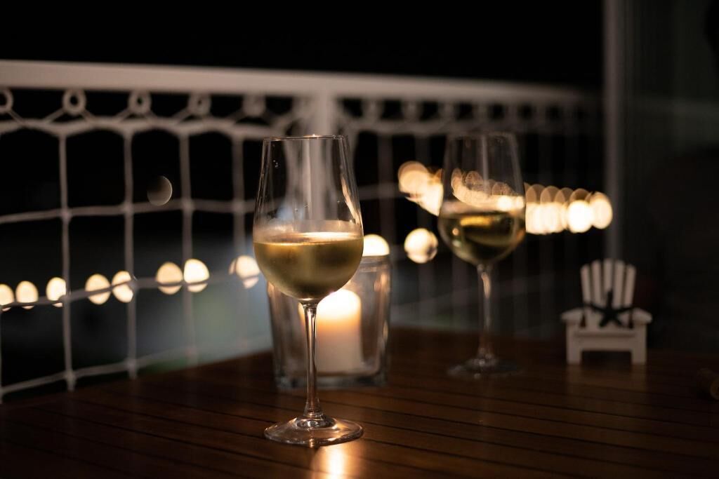 Two glasses of white wine on a wooden table with a railing and candles in the background.