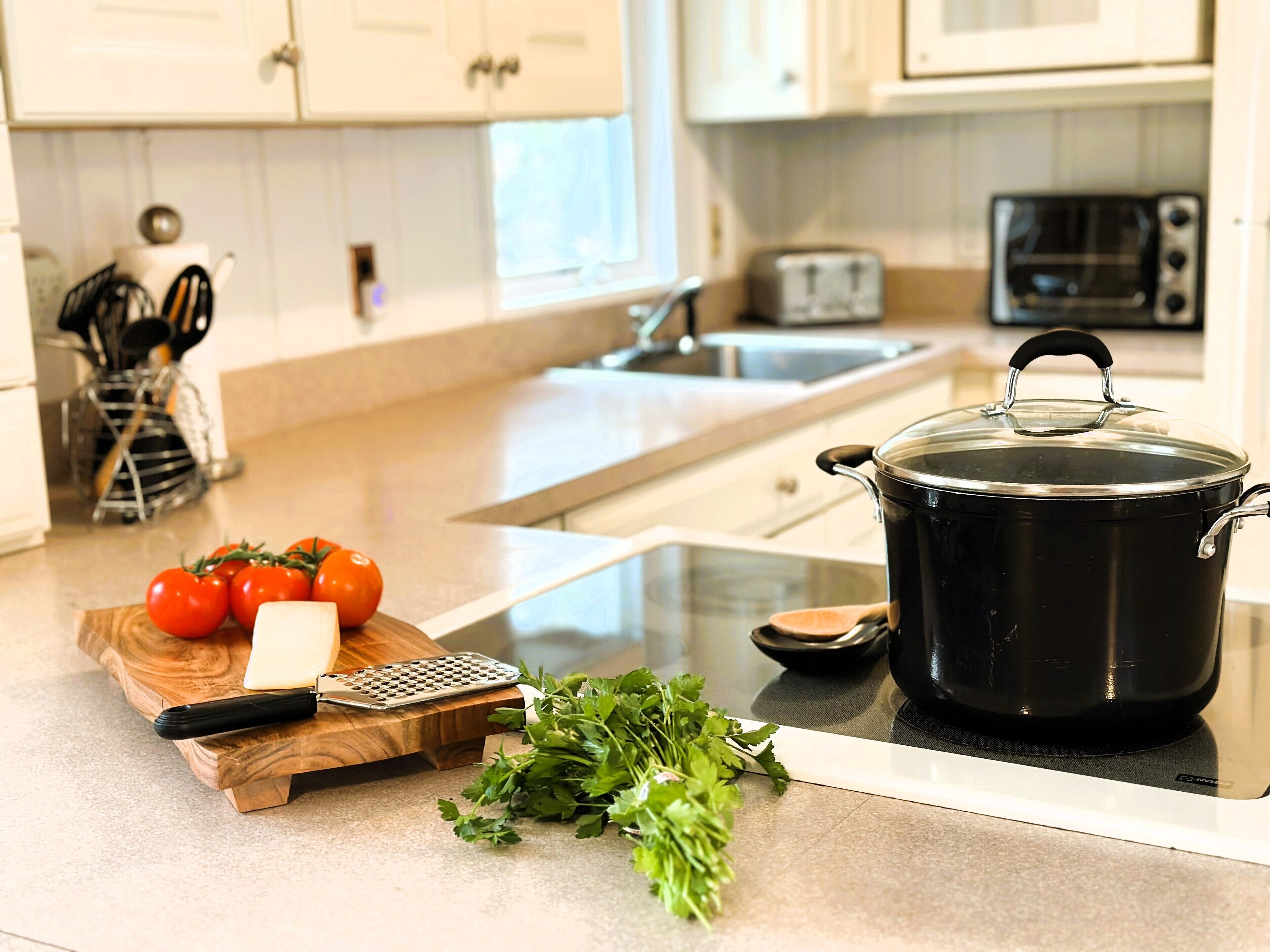 A kitchen with a stove, a pot, a cutting board with tomatoes and cheese, and a bunch of herbs.