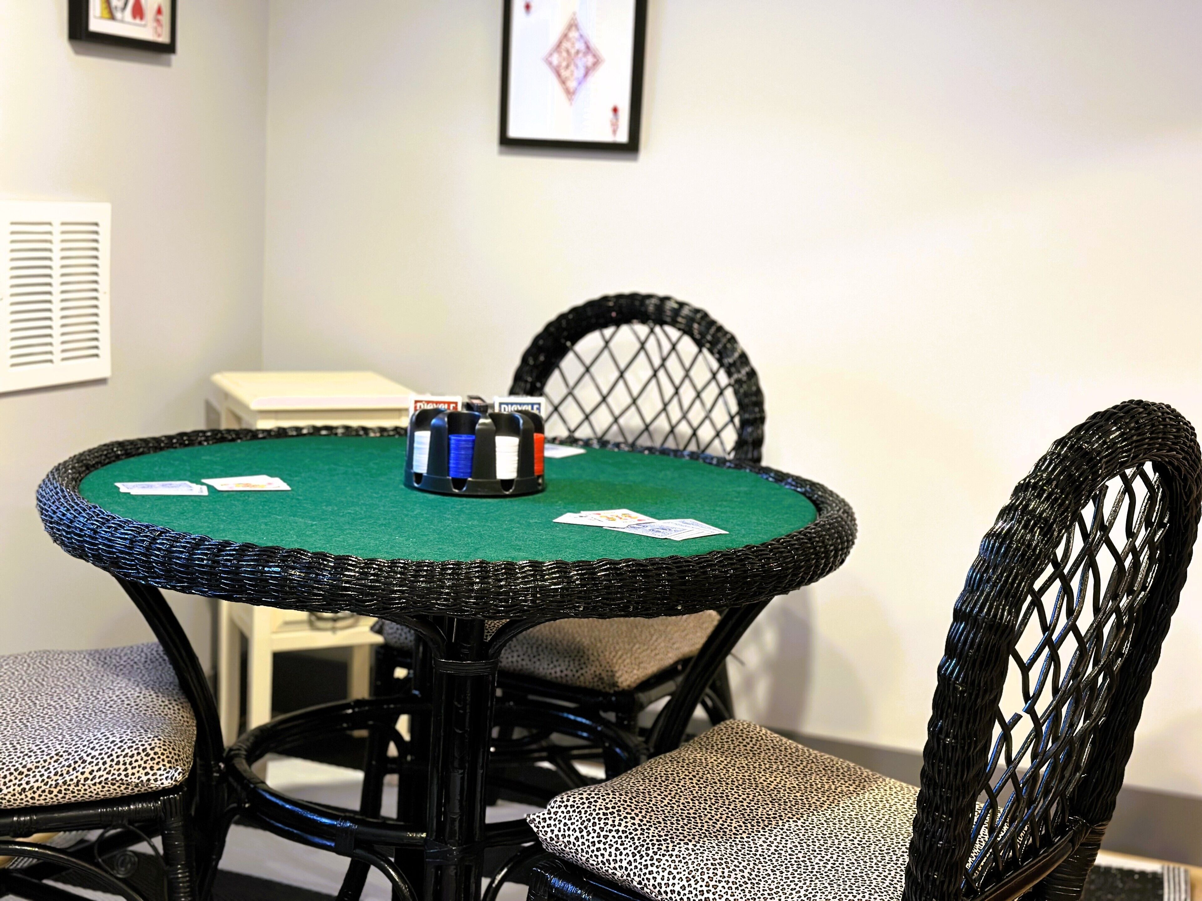 Une table ronde recouverte d’un drap de feutre vert, entourée de quatre chaises dont les dossiers sont en osier noir et les assises présentent un motif décoratif.