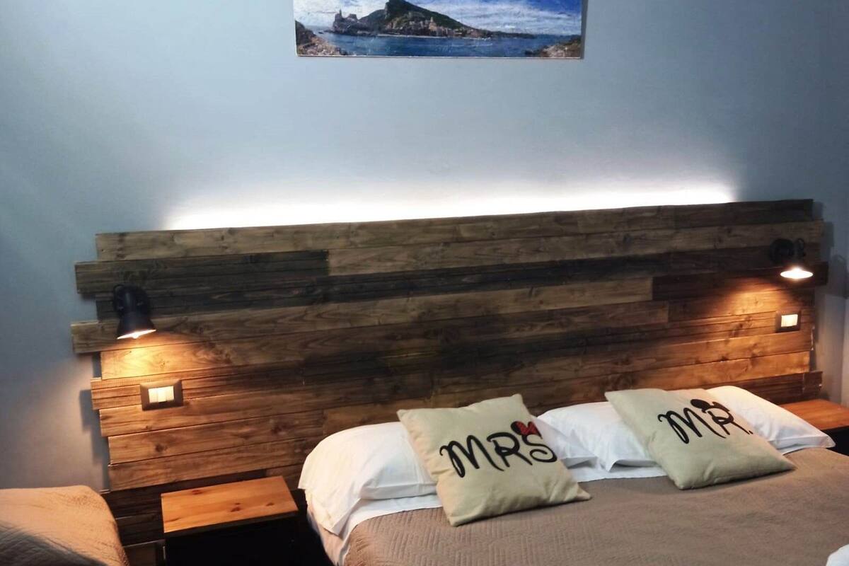 A bedroom with a wooden headboard, two bedside tables, a bed with white bedding, and two pillows with embroidered text.