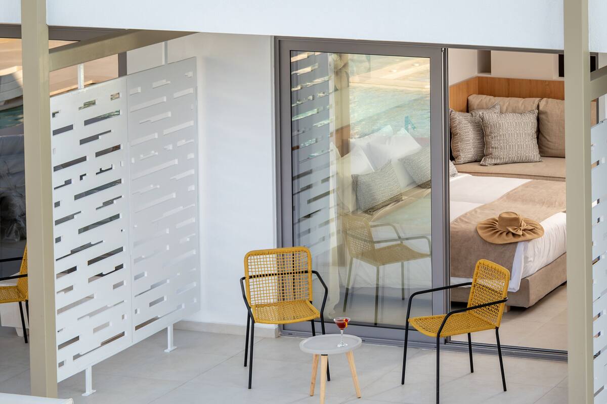 A modern outdoor lounge area with white sofas, yellow wicker chairs, and a glass-enclosed bedroom with a bed and pillows.