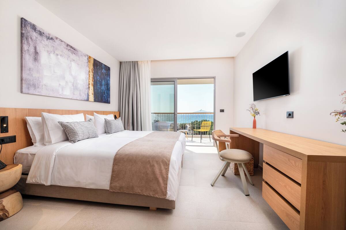 A modern hotel room with a large bed, a desk, a TV, and a balcony with a city view.