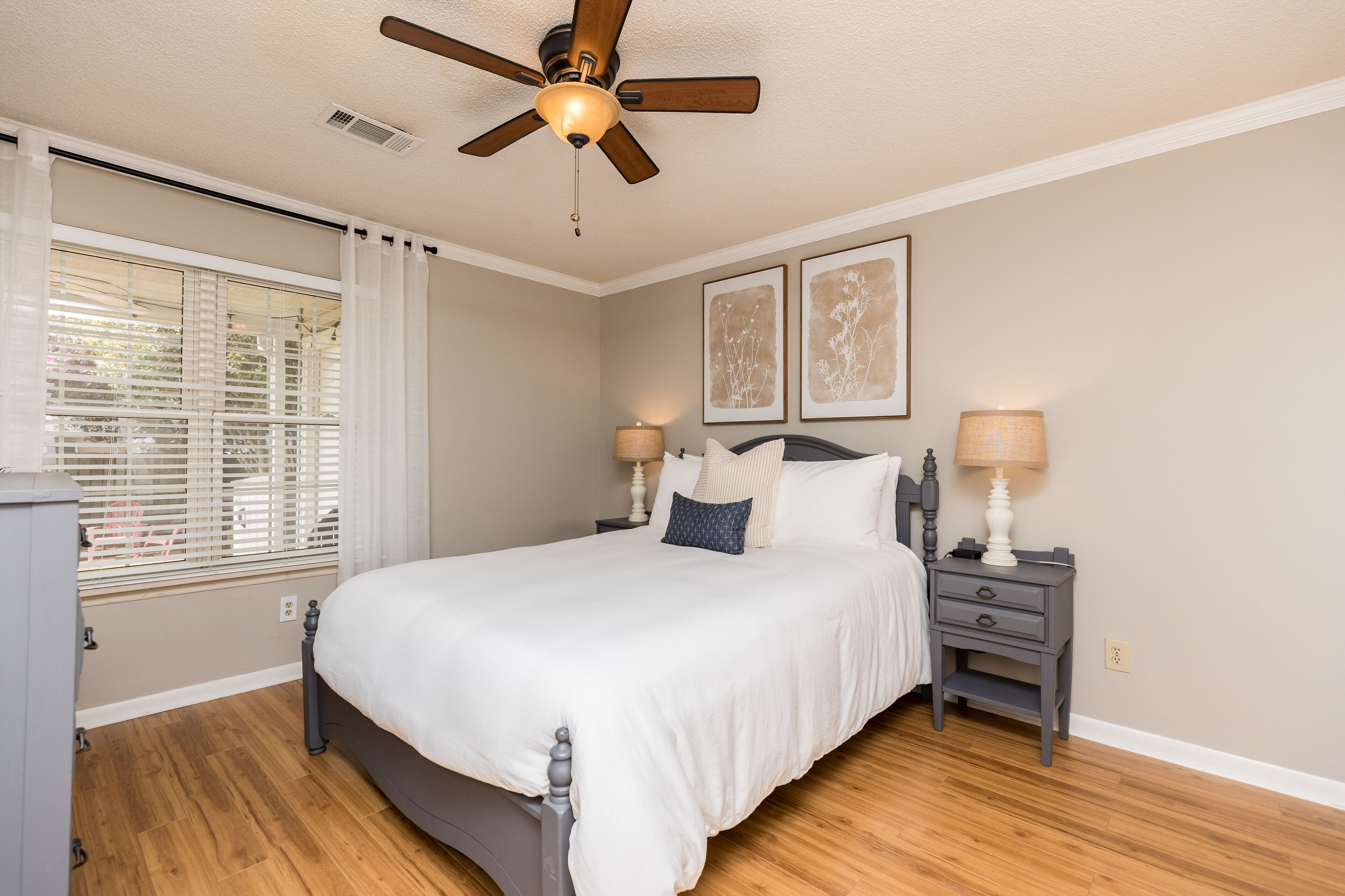 Une chambre à coucher comprenant un lit, deux tables de chevet, un ventilateur de plafond et une fenêtre avec des stores.
