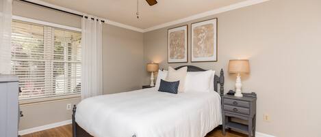 Une chambre à coucher comprenant un lit, deux tables de chevet, un ventilateur de plafond et une fenêtre avec des stores.