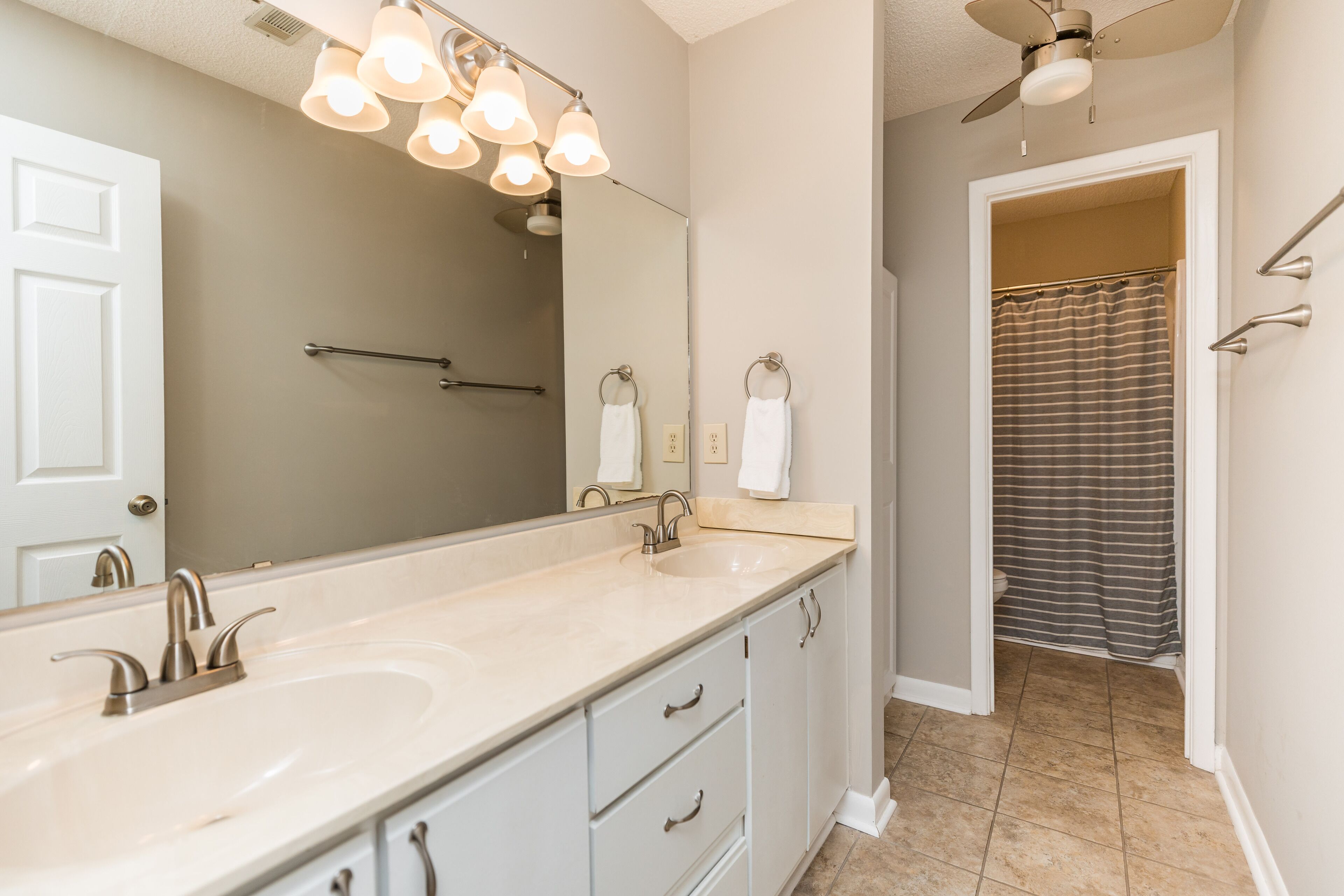 Une salle de bain avec un double lavabo, un grand miroir, un ventilateur de plafond et un rideau de douche.