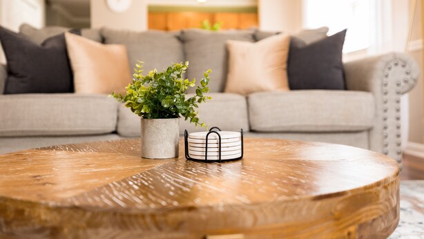 A round wooden coffee table with a potted plant and a tray in the foreground, a grey sofa with cushions in the background. - Central 2BR/2BA | Pet Friendly | Fenced Yard (Warner Robins)