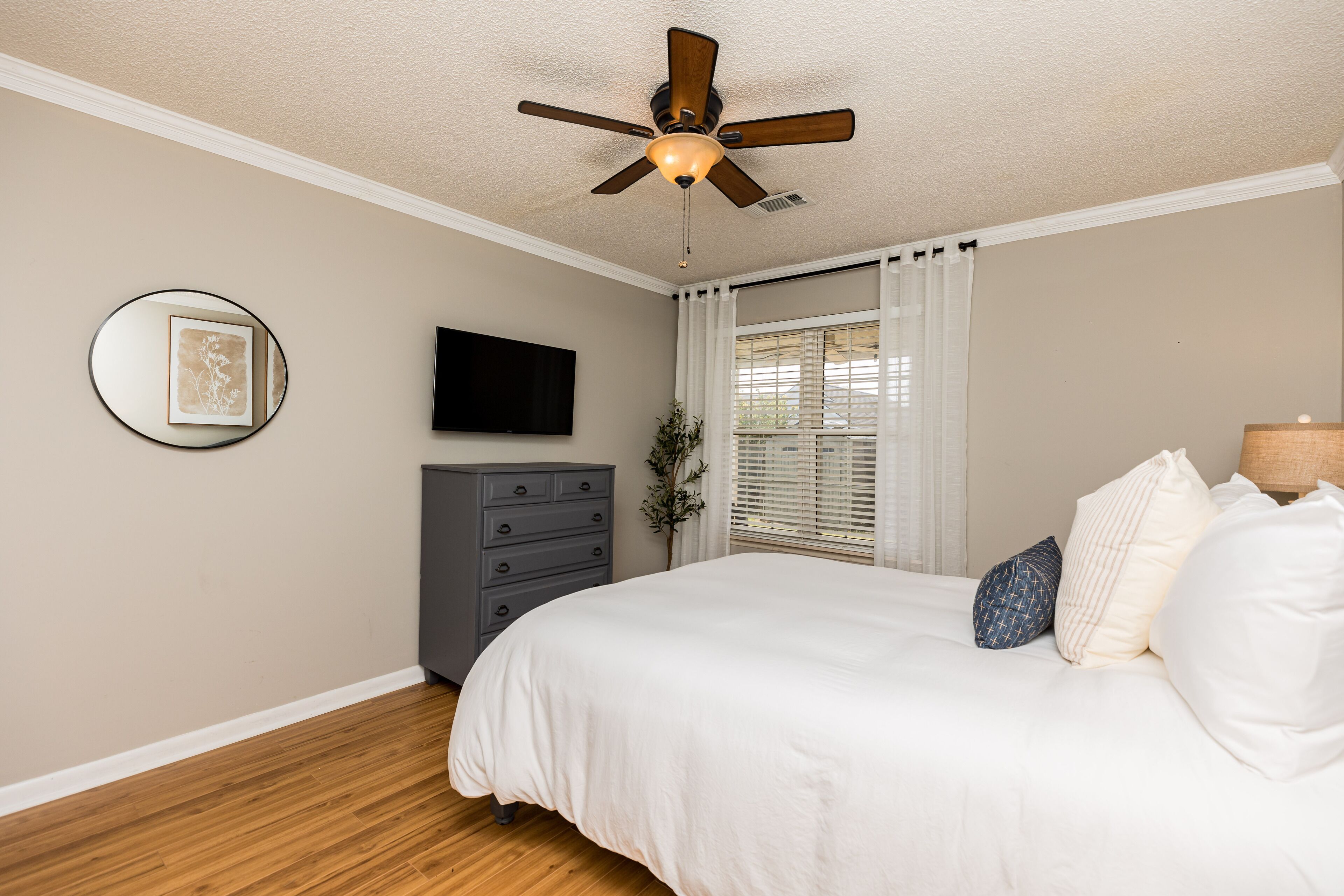 A bedroom with a bed, a ceiling fan, a dresser, a TV, and a window with curtains.