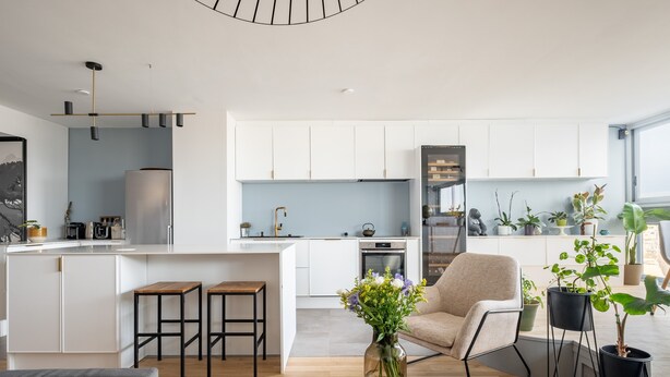 A modern kitchen with white cabinets, a central island, and a dining area with a wooden table and bar stools. There are potted plants and a vase with flowers on the table.