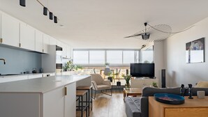 A modern apartment with a kitchen, dining area, and living room. It features a large window, a flat-screen TV, and a ceiling fan.