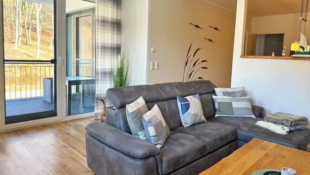 Ein modernes Wohnzimmer mit einem grauen Ecksofa, einem Holztisch und Blick auf einen Balkon mit einer Glastür.