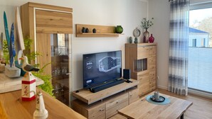 Ein modernes Wohnzimmer mit Fernseher, Holzmöbeln und einem großen Fenster mit Vorhängen.