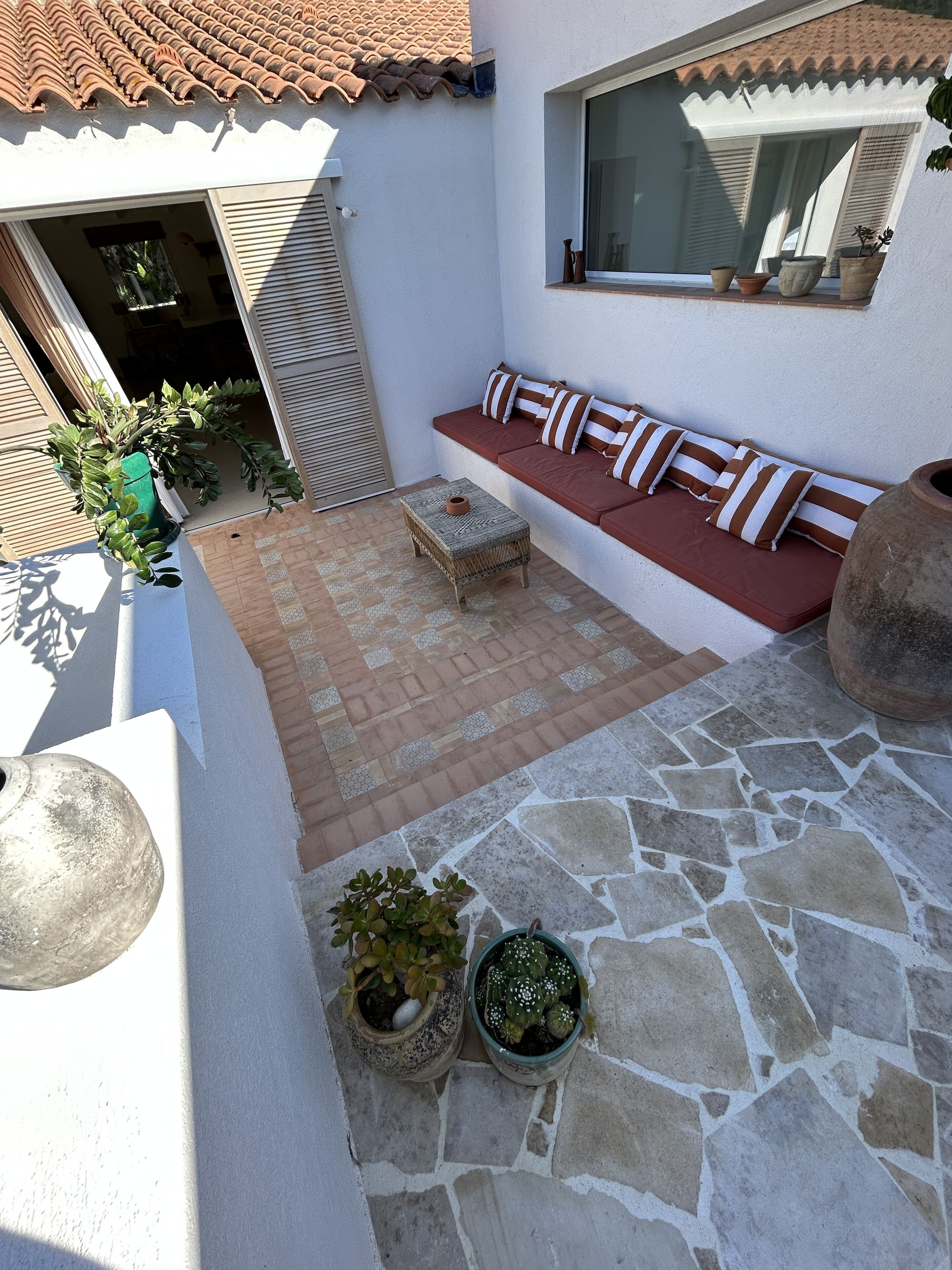 A patio area with a stone floor, a cushioned bench with striped cushions, and potted plants.