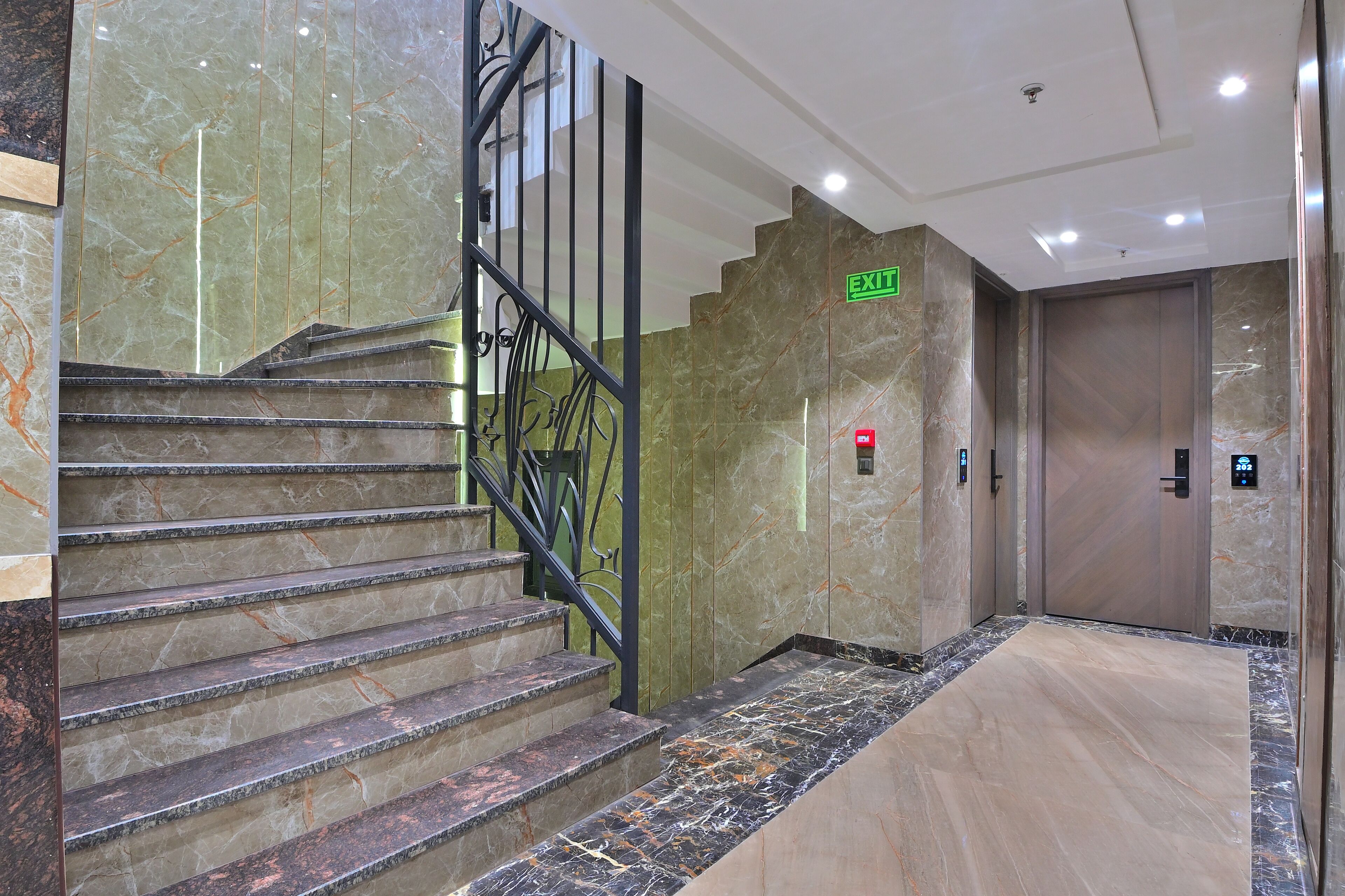 Un escalier en marbre avec un panneau indiquant la sortie, une alarme d’urgence fixée au mur et une porte donnant accès à un ascenseur.