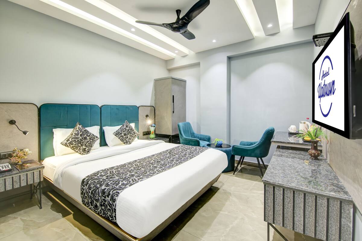 A modern hotel room with a large bed, two chairs, a desk, and a TV on the wall.