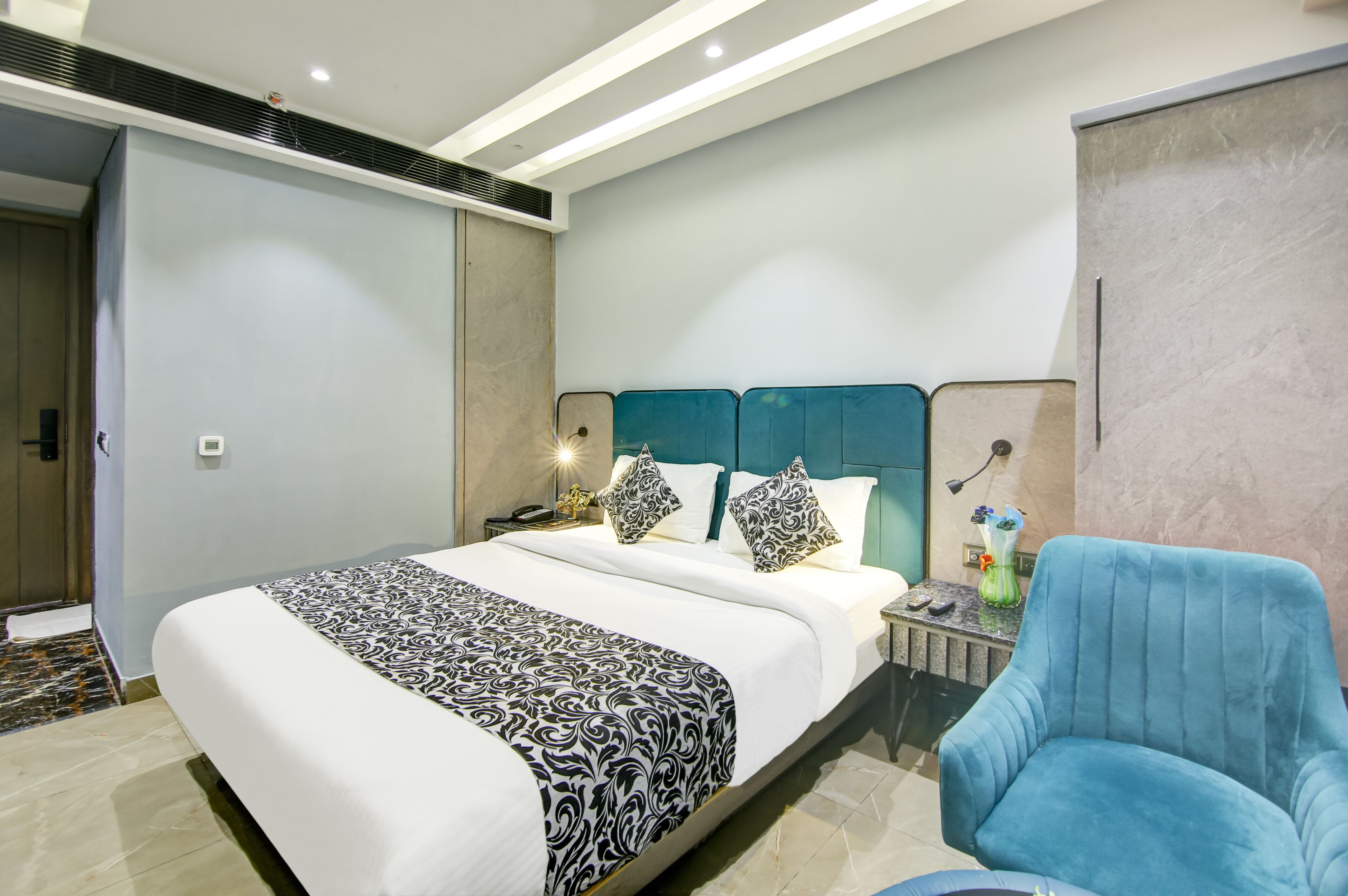 Une chambre d’hôtel moderne avec un grand lit, un fauteuil bleu, une table de chevet avec une lampe et une petite table sur laquelle est posé un vase.