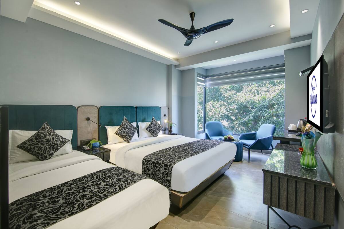 A modern hotel room with two beds, a ceiling fan, a large window with a view of trees, and a flat-screen TV on the wall.