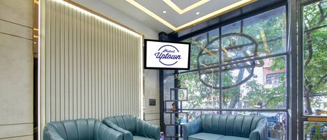 A modern hotel lobby with two sofas, a TV screen, and a large window with a view of the city.