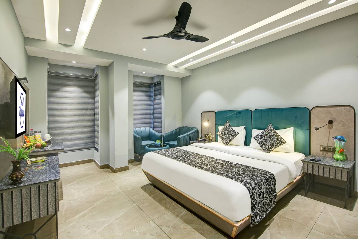 A modern hotel room with a large bed, a ceiling fan, a desk with a vase, and a seating area with a blue chair.