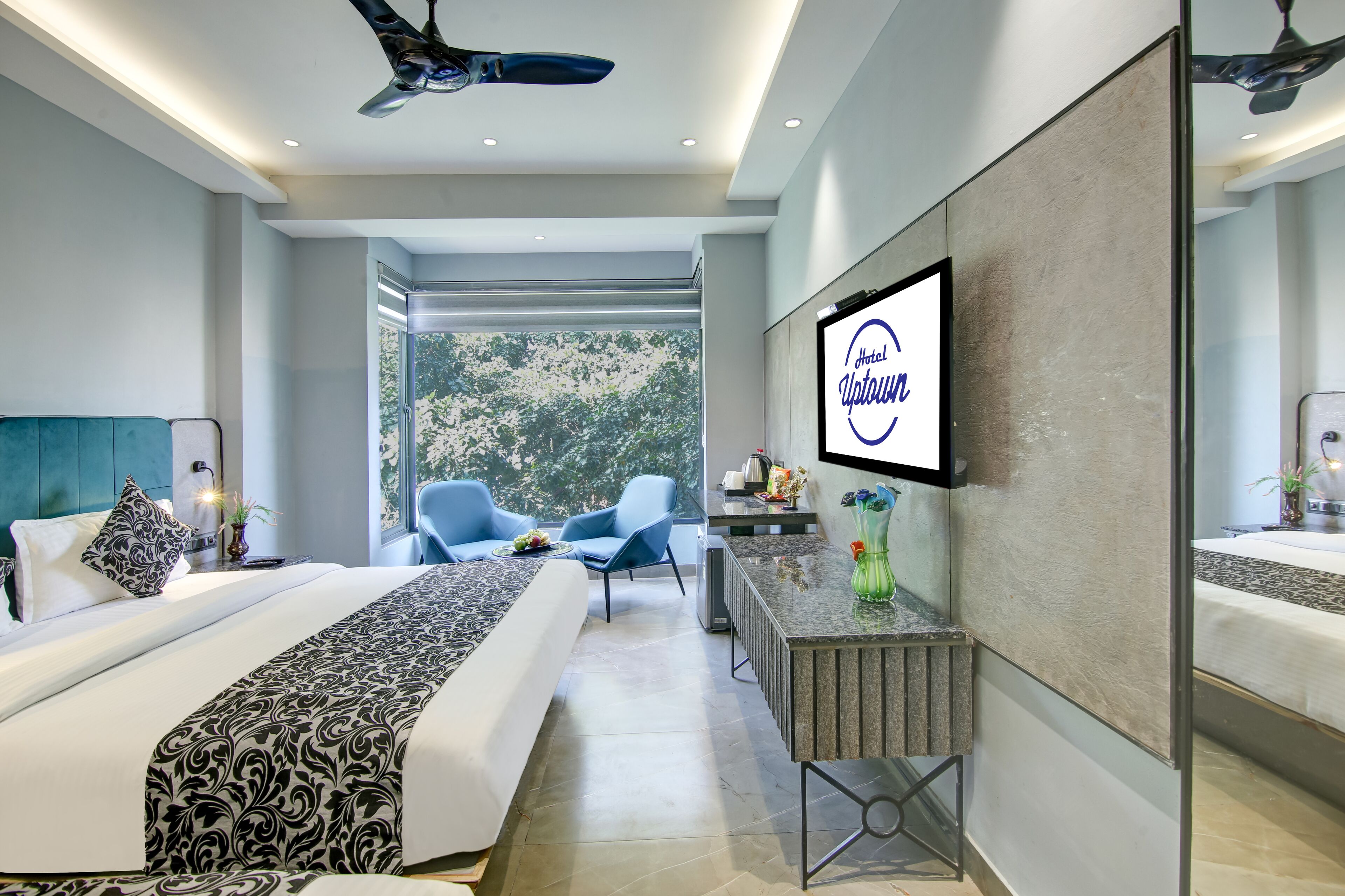A modern hotel room with a large bed, a desk, a chair, and a TV displaying the hotel name.
