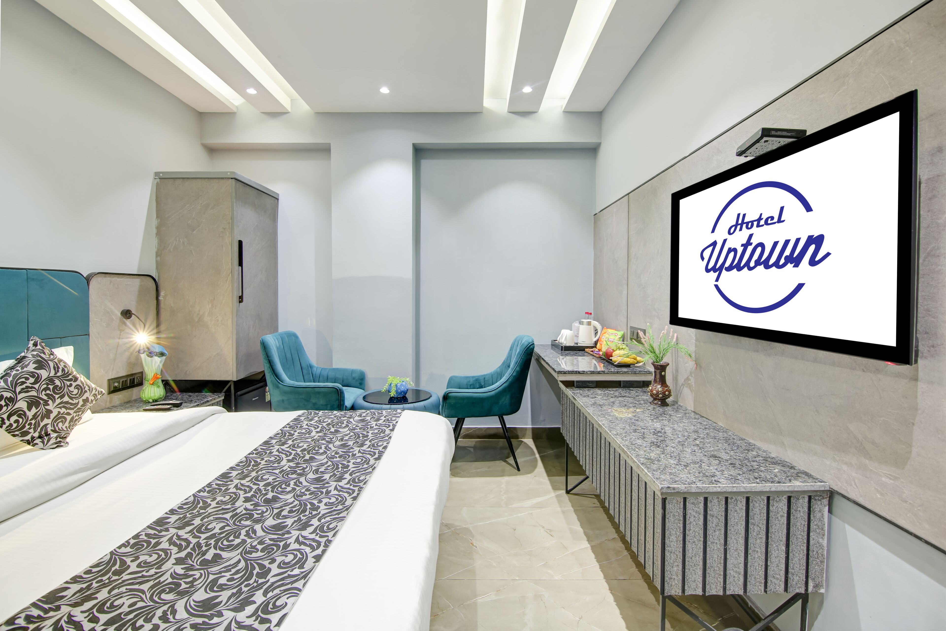 Hotellihuone, jossa on sänky, kaksi tuolia, pieni pöytä maljakolla ja seinälle asennettu televisio, jossa näkyy teksti ”Hotel Uptown”.