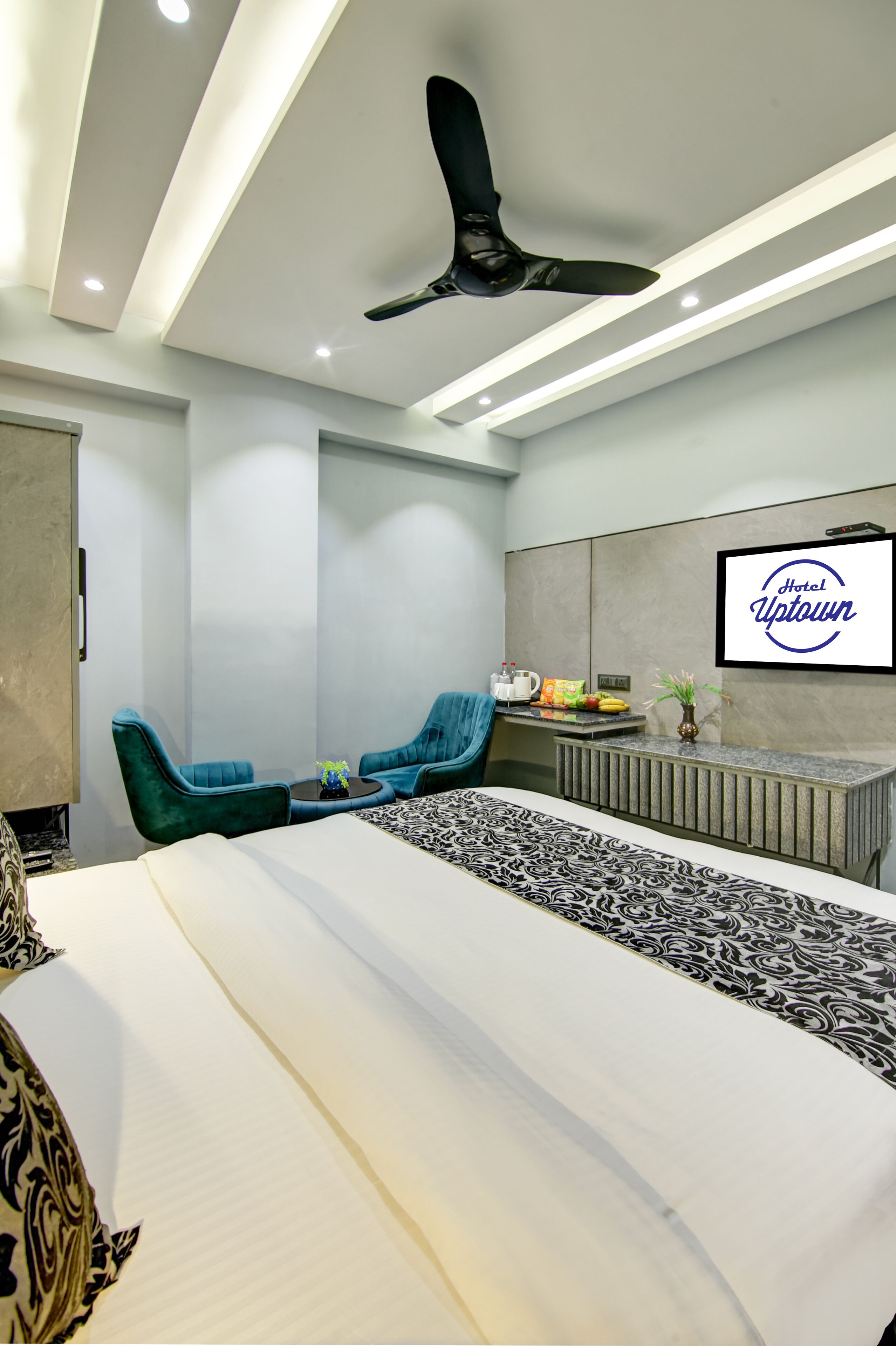 A hotel room with a bed, a seating area with blue chairs, a ceiling fan, and a wall-mounted TV displaying 'Hotel Uptown'.